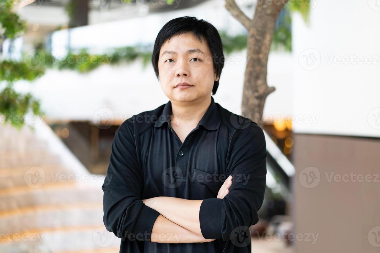The portrait of Asian man in a black casual shirt with crossed arms standing in front of a blurred background of an office workspace. photo