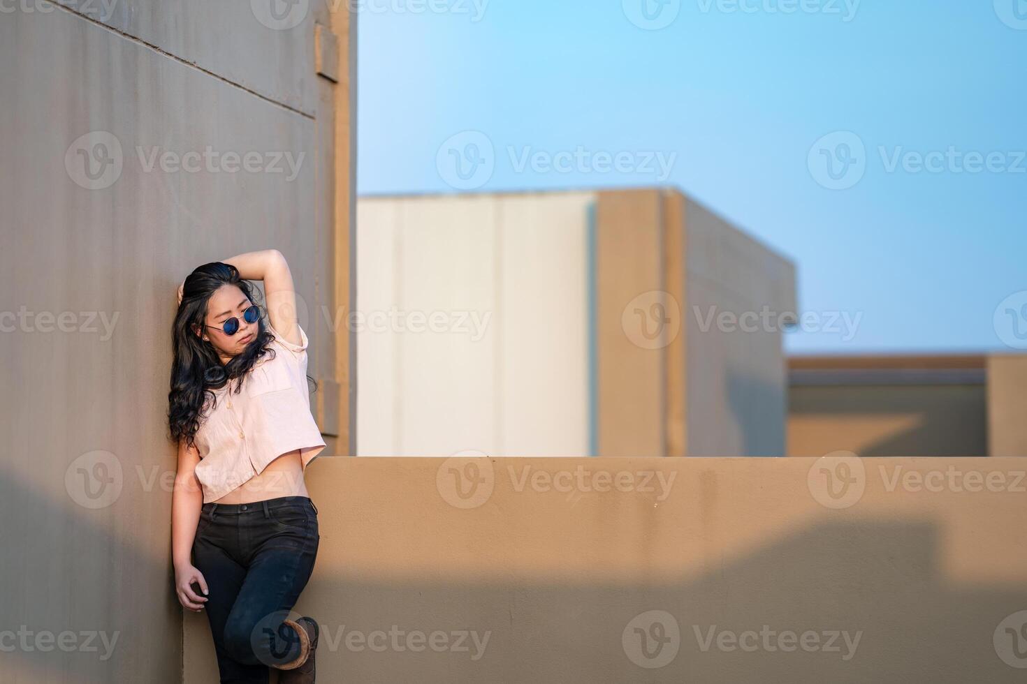 A cute Asian woman stands up and raise elbow up against the wall and wears blue sunglasses at the rooftop of the building at sunset time. photo