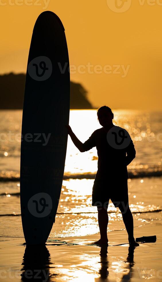 Silhouette Smart Asian Man long hair touches and poses with Standed Surfboard on the beach in Twilight time photo