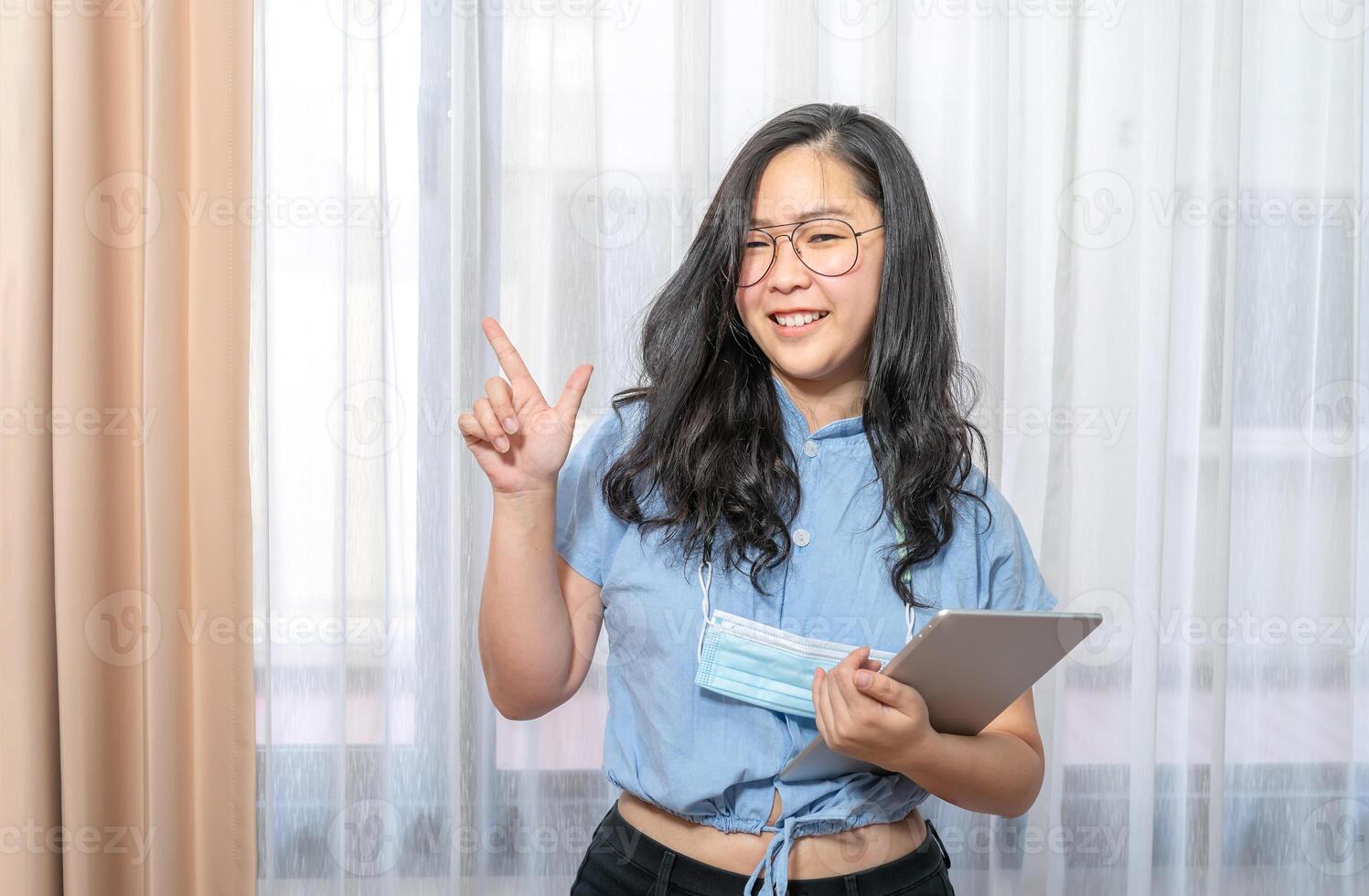 Happy Asian woman fingering up and pointing with tablet computer on her hand at copy space while standing in front of the transparent curtain see through the window light. photo