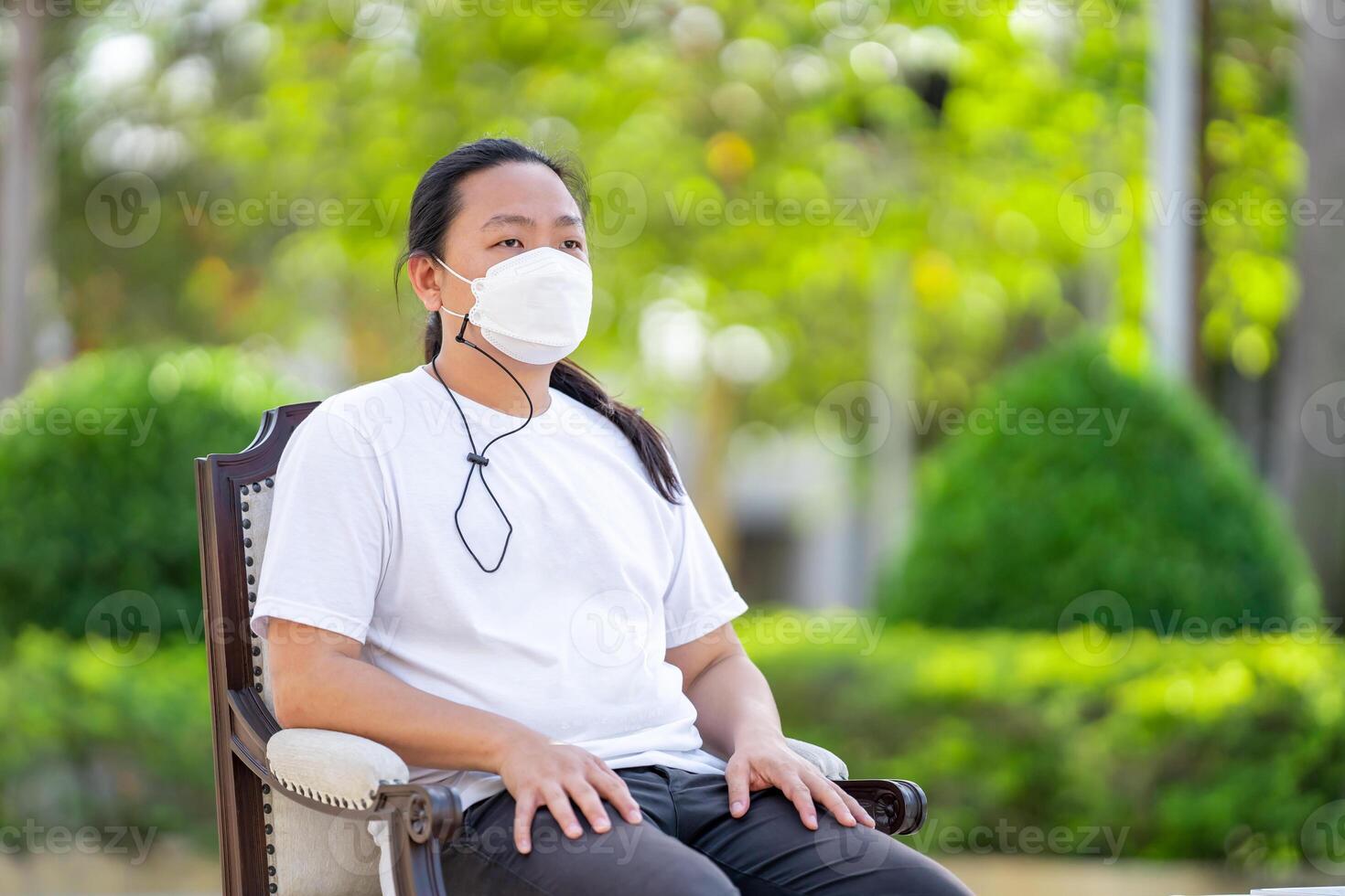 A man wearing white T shirt and white mask is sitting calm on the armchair with his arms on the armrest and his back on the backrest. photo