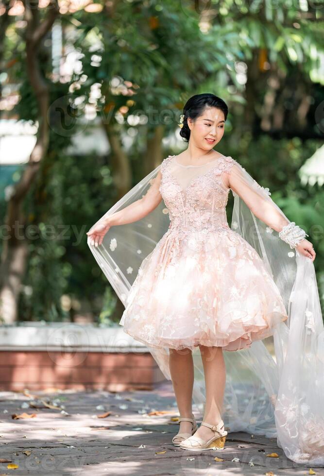 Asian girl in an old rose pink color knee length fluffy dress with high heel slingback shoes and ceremonial thread on her wrist. She is brighter mood posing in the shade of tree garden. photo