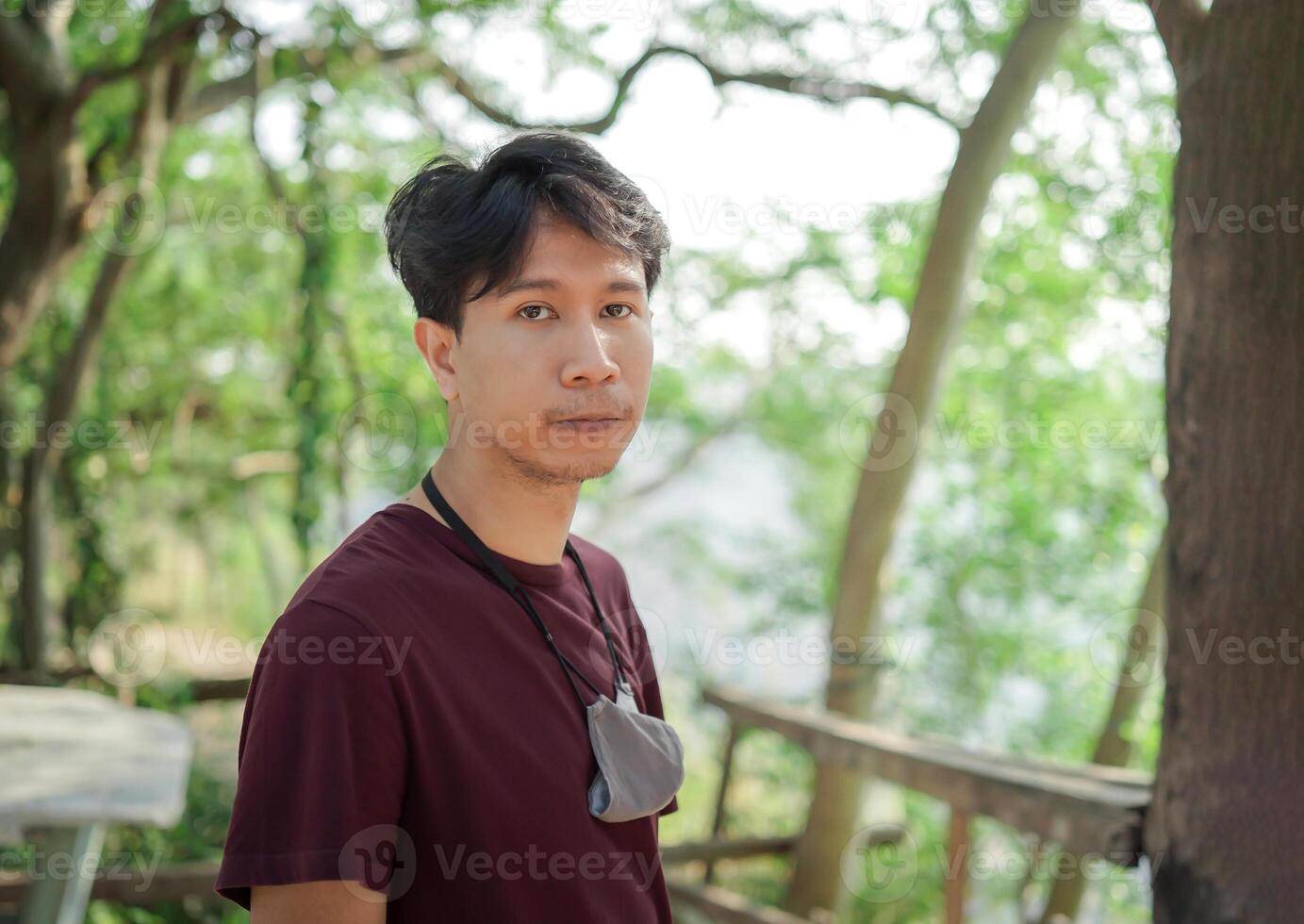 Asian Smart man in casual dark red T-shirt and hang on the gray cloth mask on his neck, stands and looks to the camera with the green tree blur background. photo