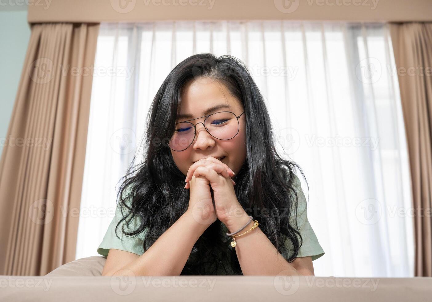 Beautiful Asian woman hold her hands together while lie on her stomach on bed in bedroom with a pastel green - brown color theme. photo