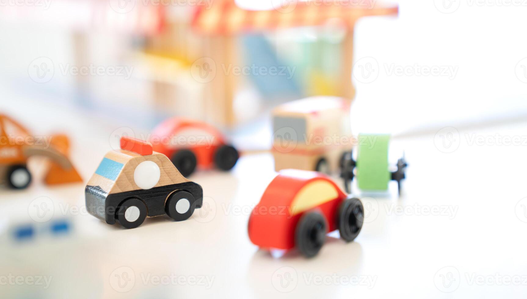 In an indoor playground, a wooden Police car toy drives behind in an Ambulance Van with a variety of cars trailing around it. photo
