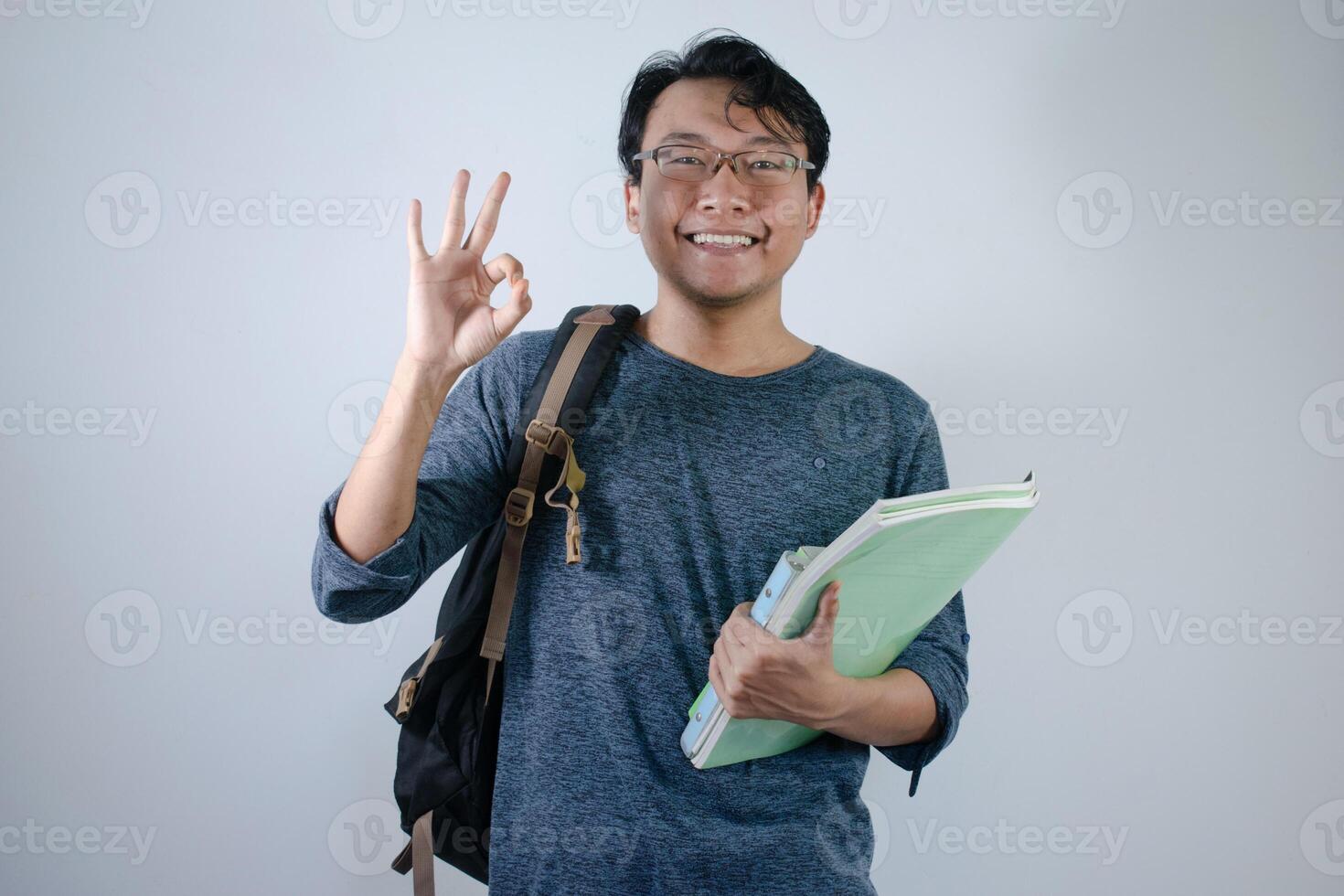 Asian young male student wearing bag and holding book smiling and happy expression. Ok hand sign gesture with isolated white background photo