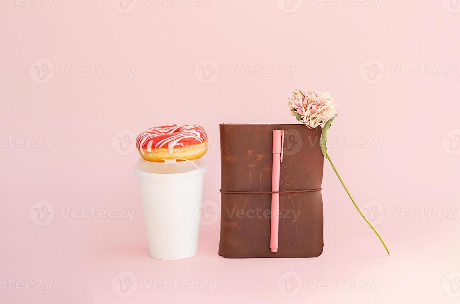 Donut on top of disposable coffee cup, notebook and dried flower on pink background. photo