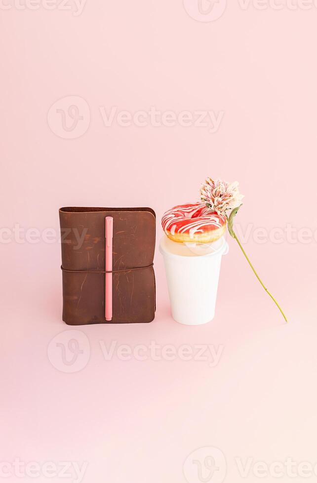 Donut on top of disposable coffee cup, notebook and dried flower on pink background. photo