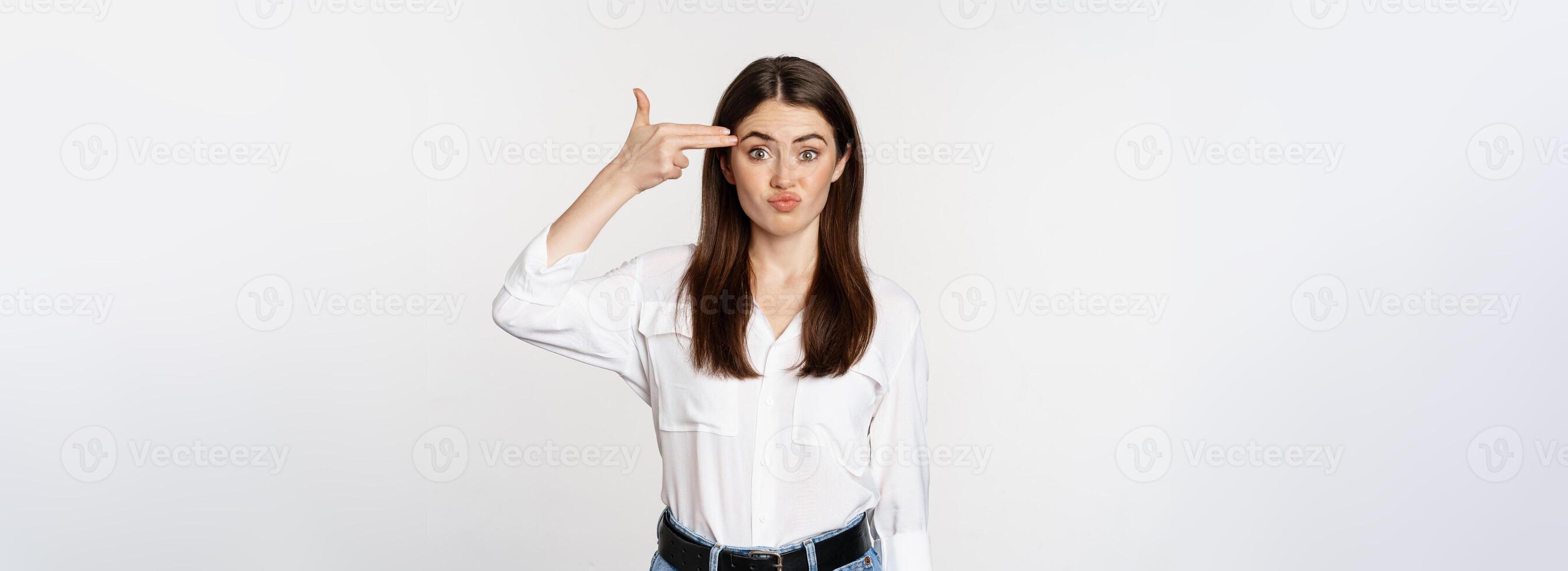 Image of annoyed young woman showing finger gun gesture over head, bothered by smth, tired, standing over white background photo