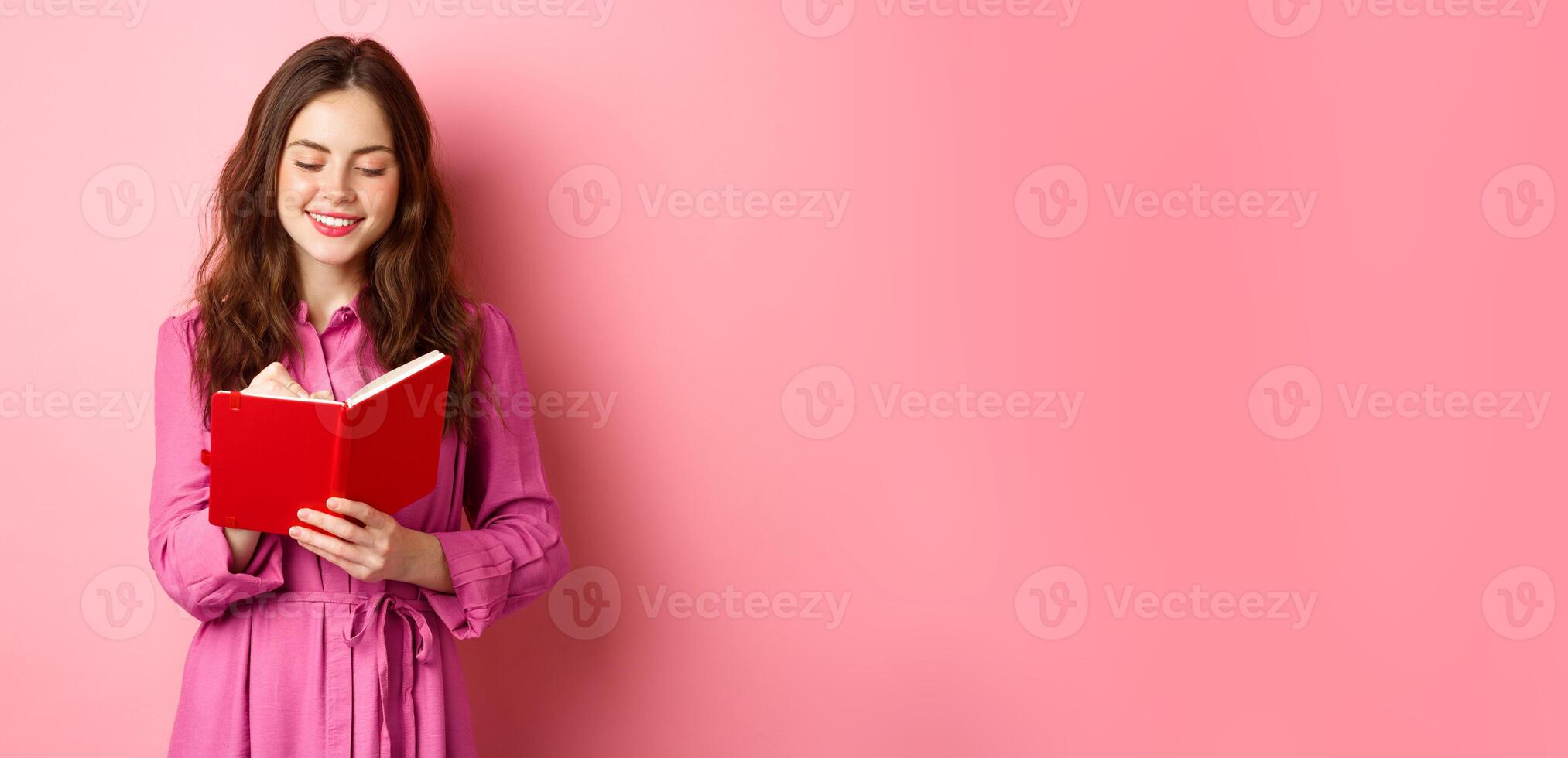 Attractive modern woman writing in her diary, making notes in notebook or planner, looking at her schedule, standing against pink background photo