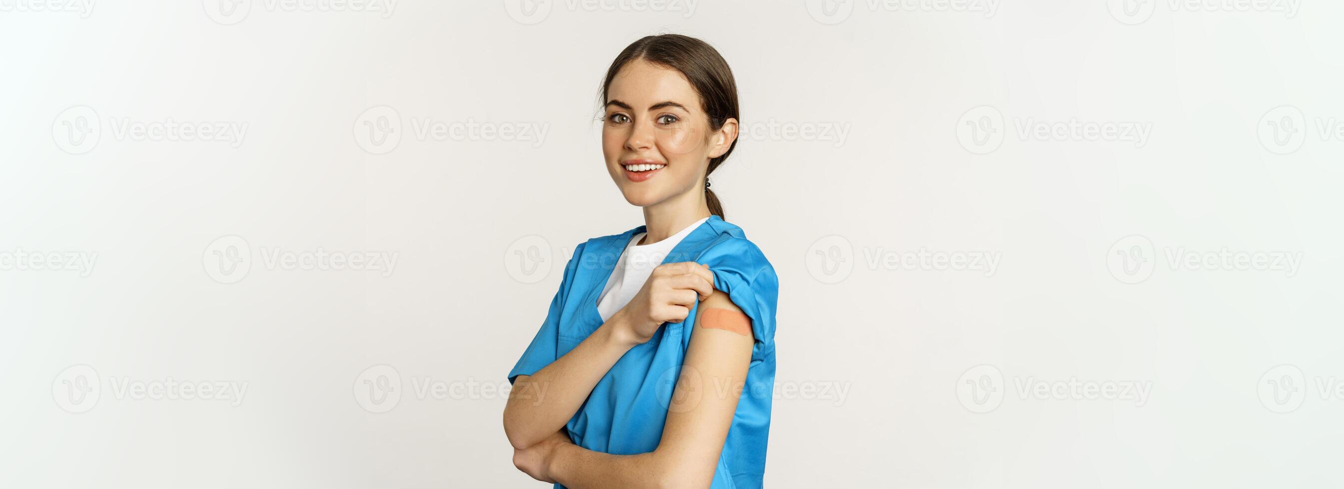 Female nurse, doctor medical worker got vaccinated, showing shoulder with patch after covid-19 vaccine, smiling pleased, standing over white background photo