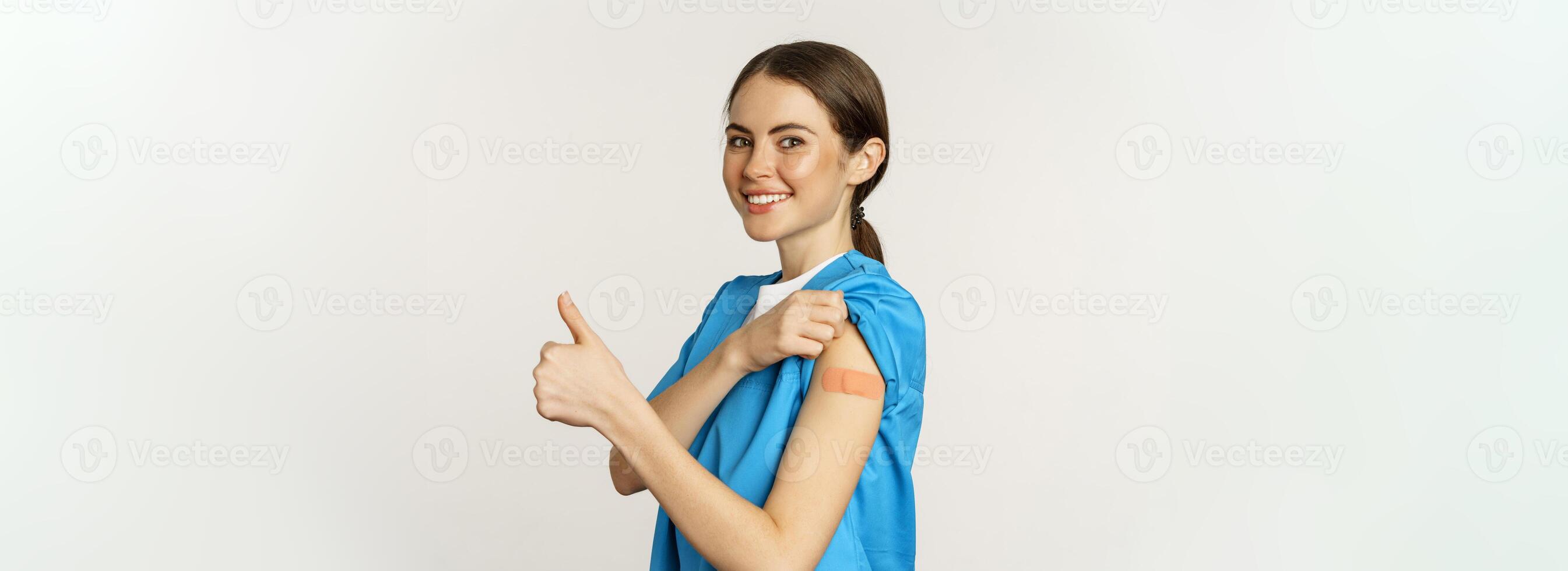 Smiling nurse, doctor in scrubs, medical worker showing her vaccinated shoulder with patch, thumbs up, recommending vaccinate from coronavirus, white background photo