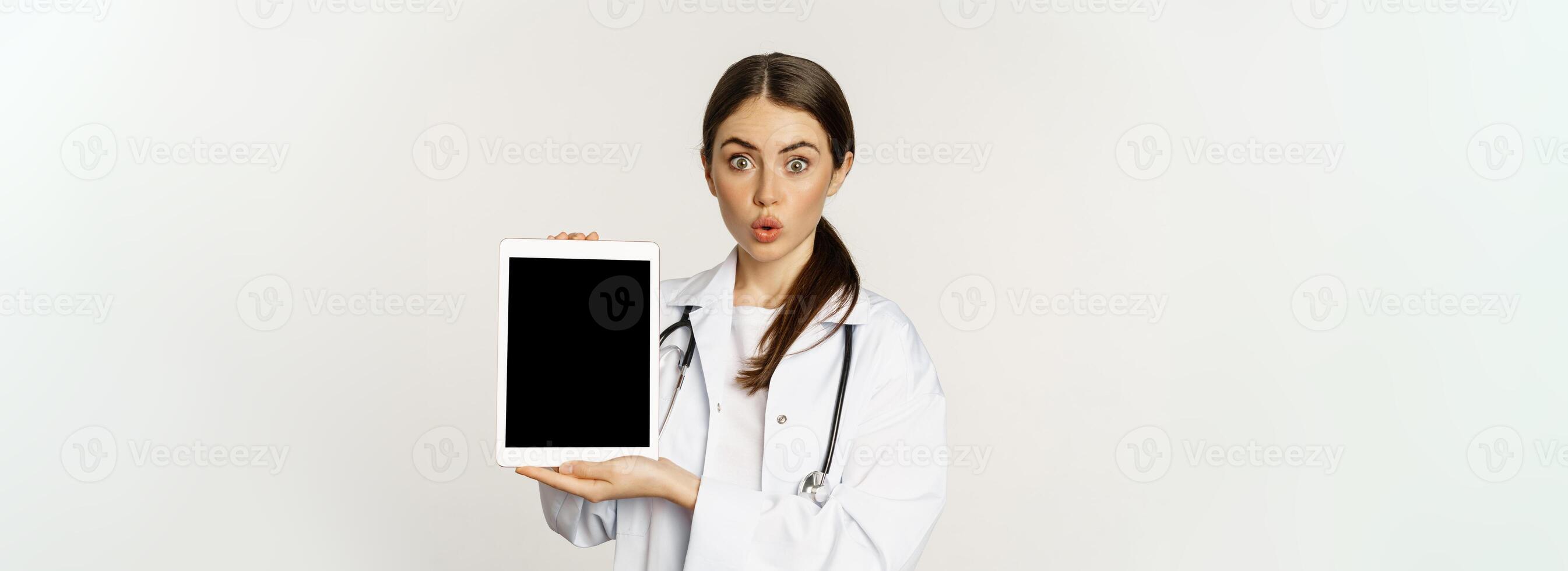 Image of woman doctor, female healthcare worker showing online medical website, digital tablet screen and smiling, standing in white coat over white background photo