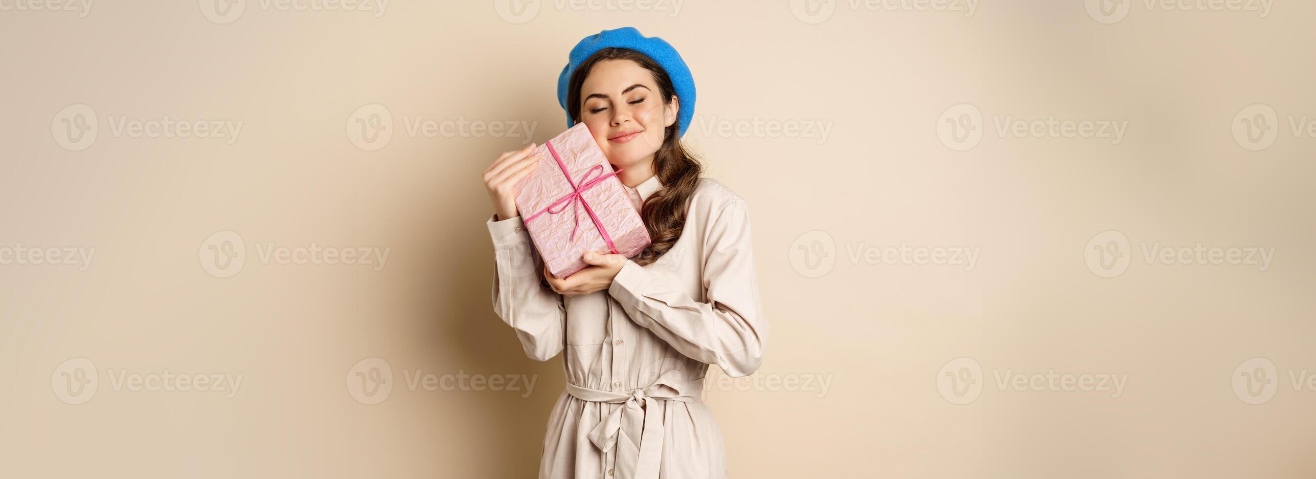 Holidays and gifts concept. Beautiful girl receive gift box and looking happy, holding pink wrapped present with joyful face expression, beige background photo