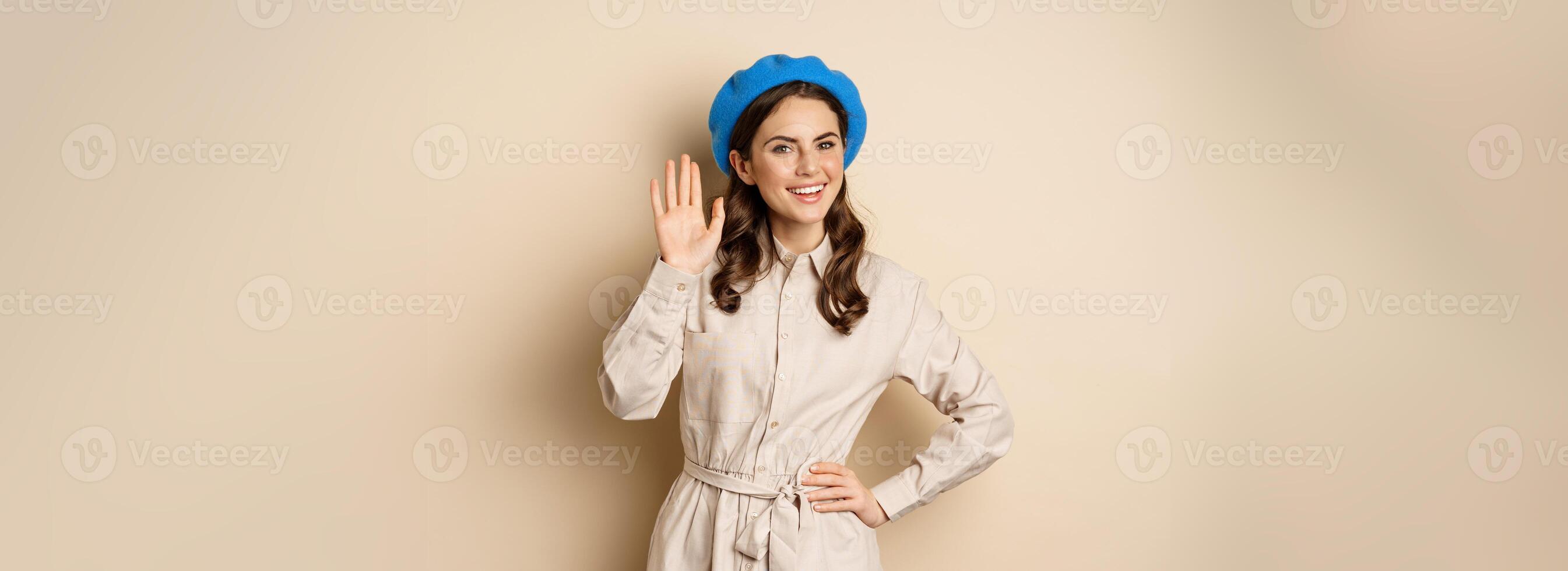 Image of beautiful friendly woman in outerwear, saying hello, waving hand and smiling, greeting you, standing over beige background photo