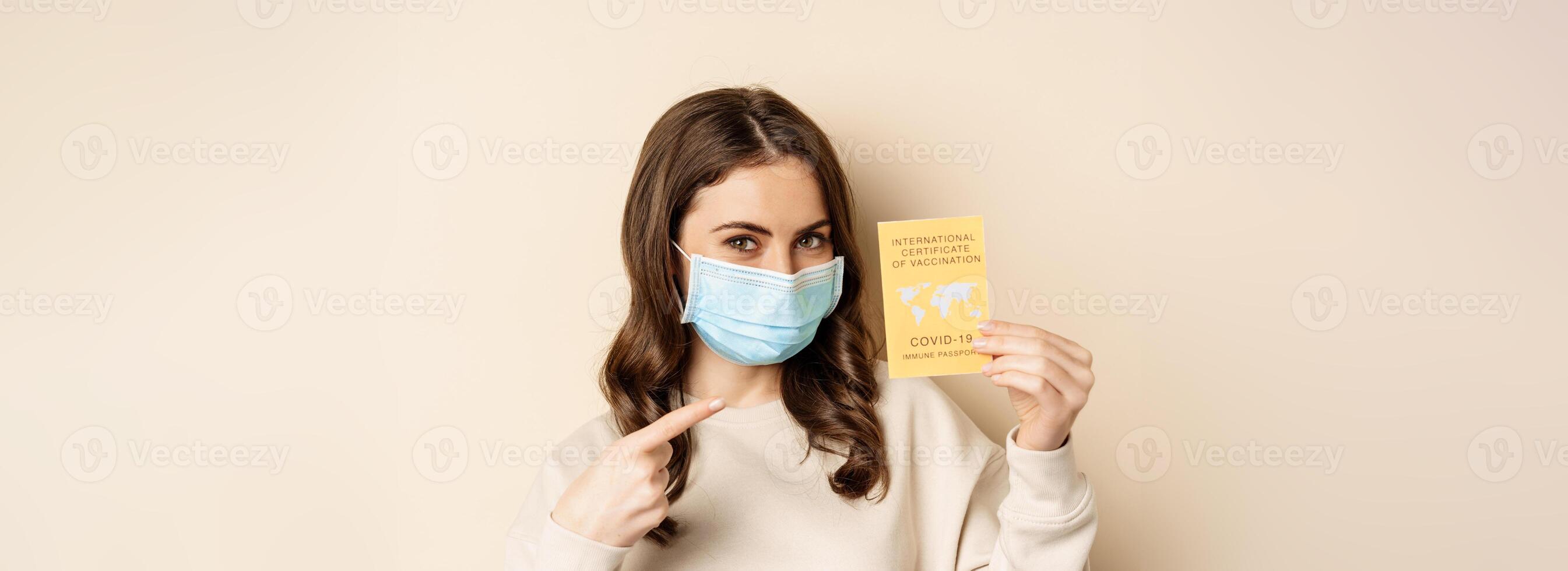 Covid and people concept. Vaccinated woman in face mask showing covid international vaccination certificate, recommend get vaccine, standing over beige background photo