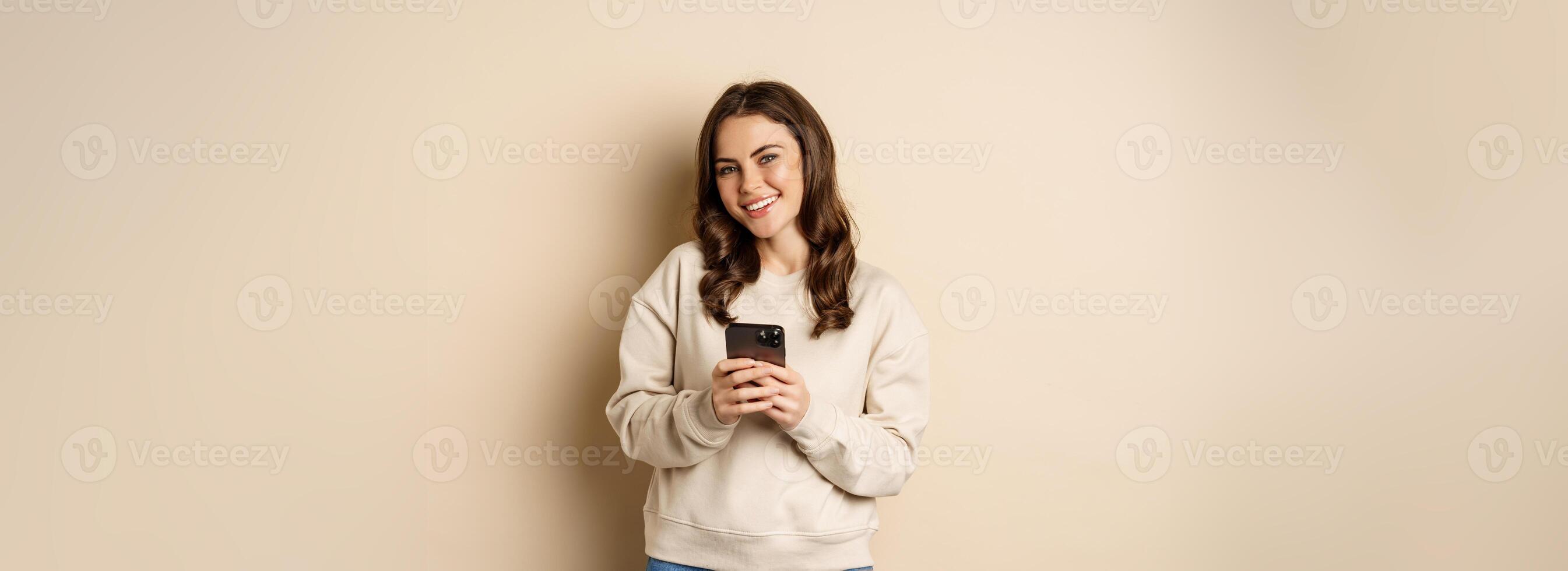 People and cellular technology. Beautiful stylish woman using mobile phone, smartphone app, smiling and looking at screen, standing over beige background photo