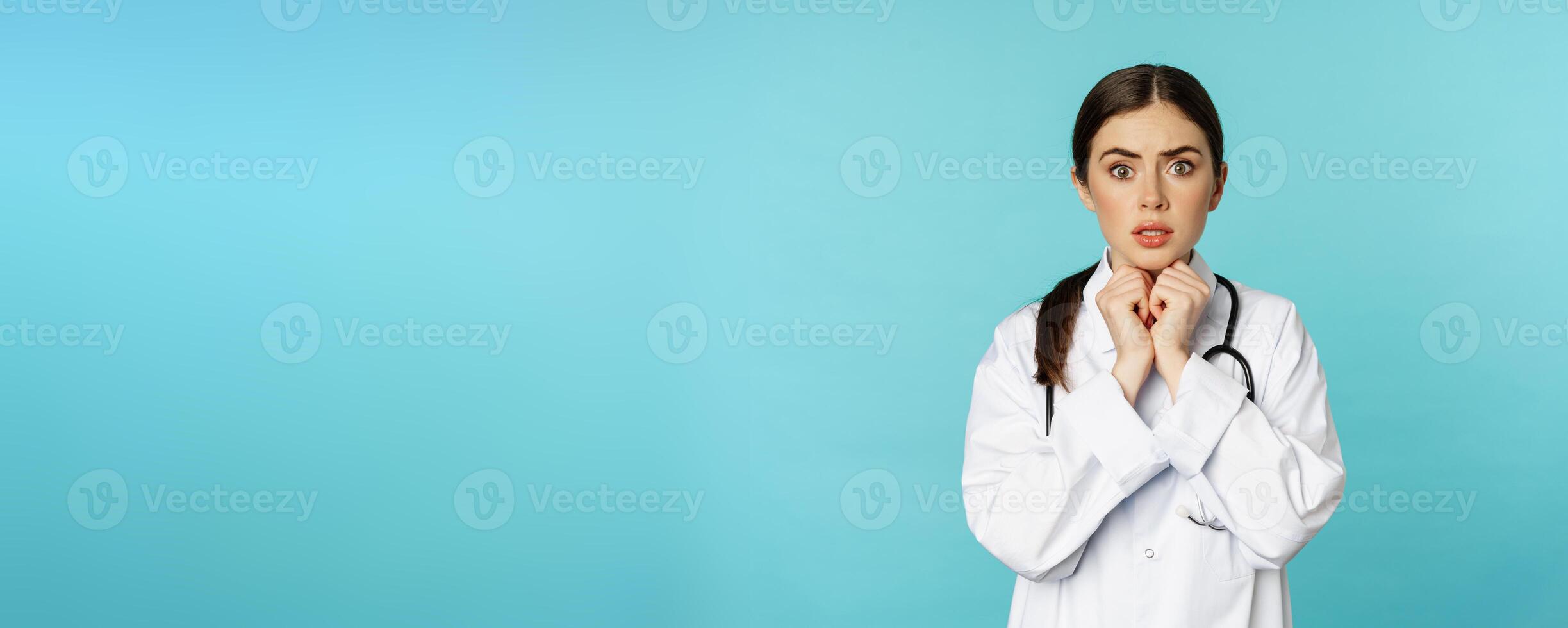 Worried and concerned physician, hospital worker in white lab coat, looking scared and nervous at camera, standing with stethoscope over torquoise background photo