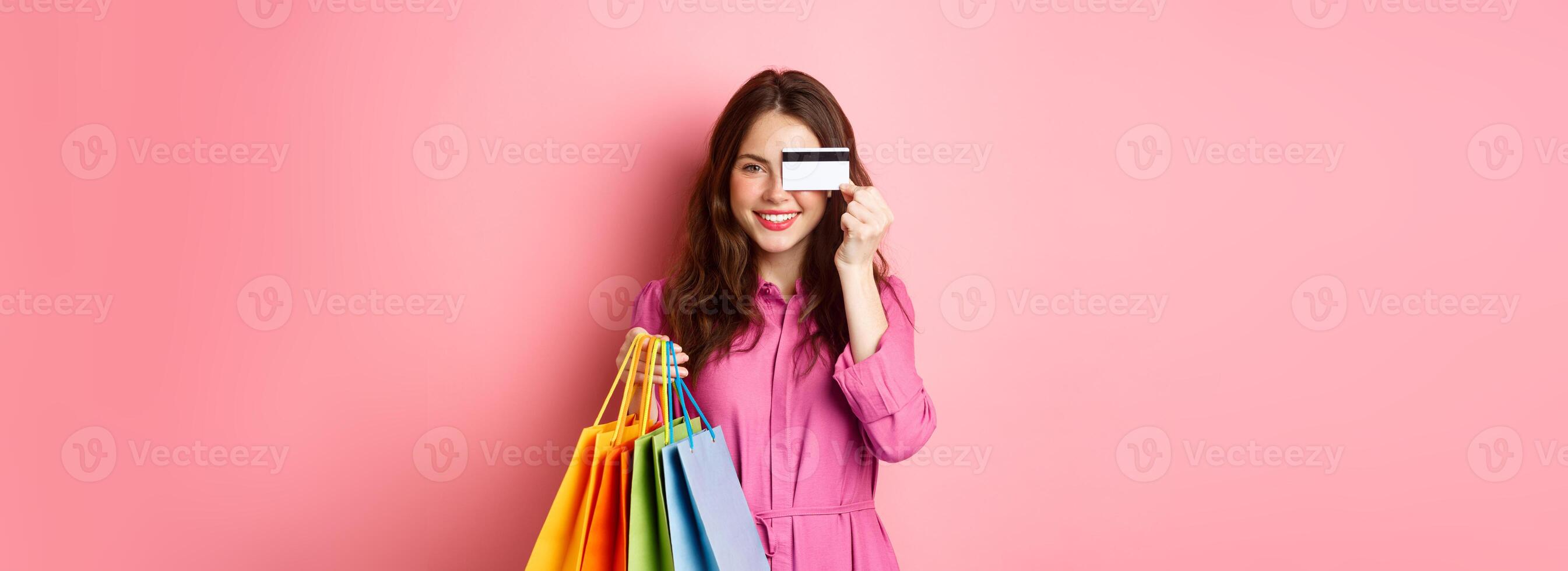 Portrait of excited happy shopaholic, woman holding shopping bags and showing plastic credit card, smiling amazed, standing against pink background photo