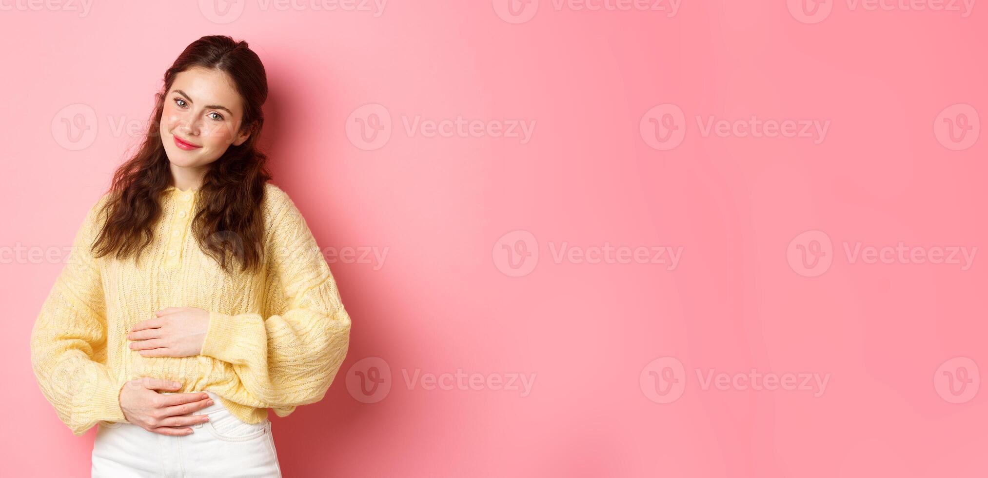 Young smiling woman being pregnant, rubbing her belly and looking with hopeful happy face at camera, expecting for baby, standing against pink background photo