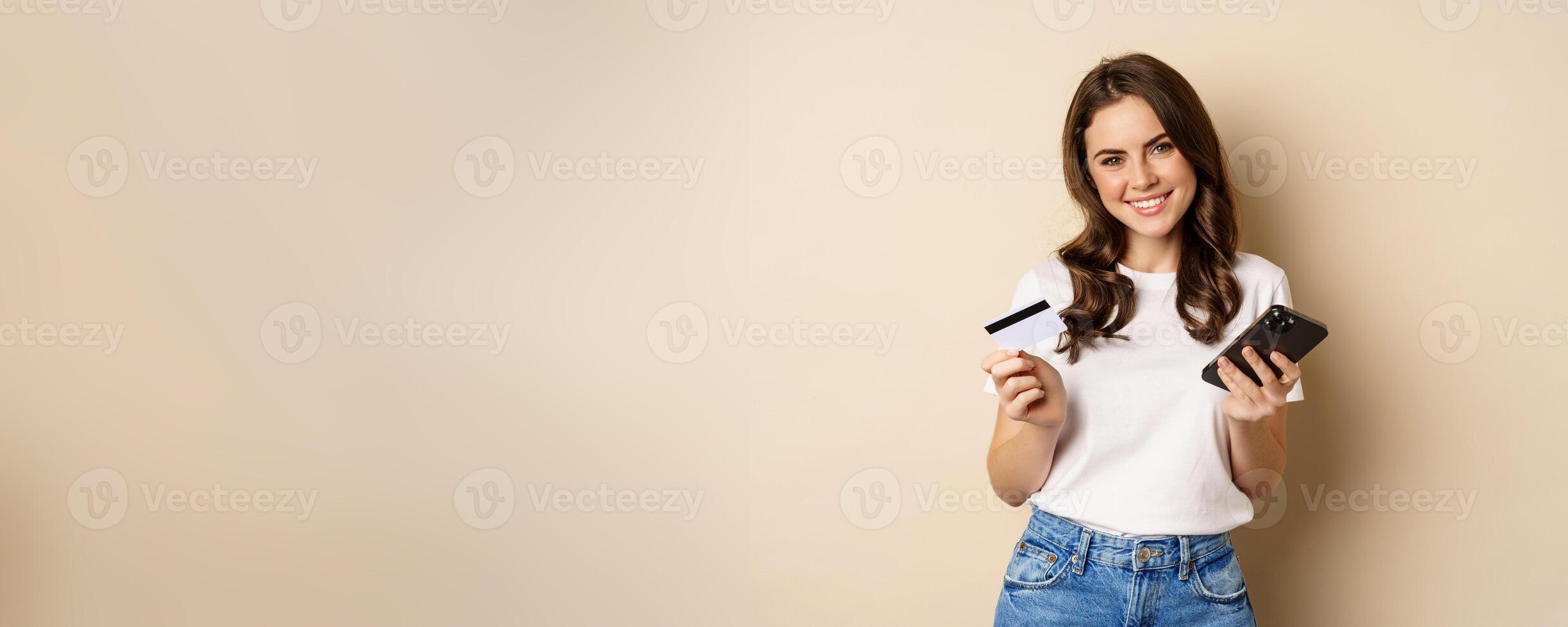 Image of young happy woman paying online, holding smartphone and credit card, enter info in application on mobile phone, standing against beige background photo