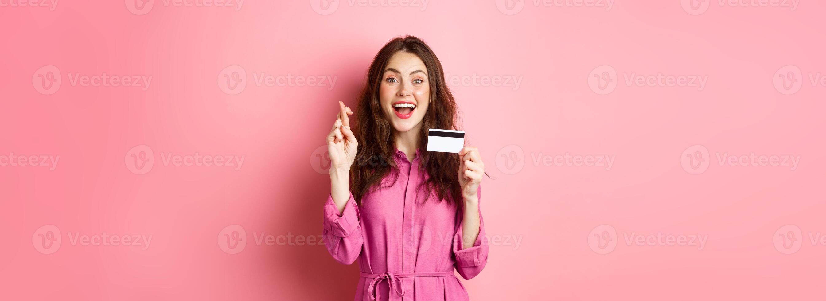 Image of hopeful young woman showing plastic credit card and cross fingers for good luck, making wish and smiling, standing against pink background photo