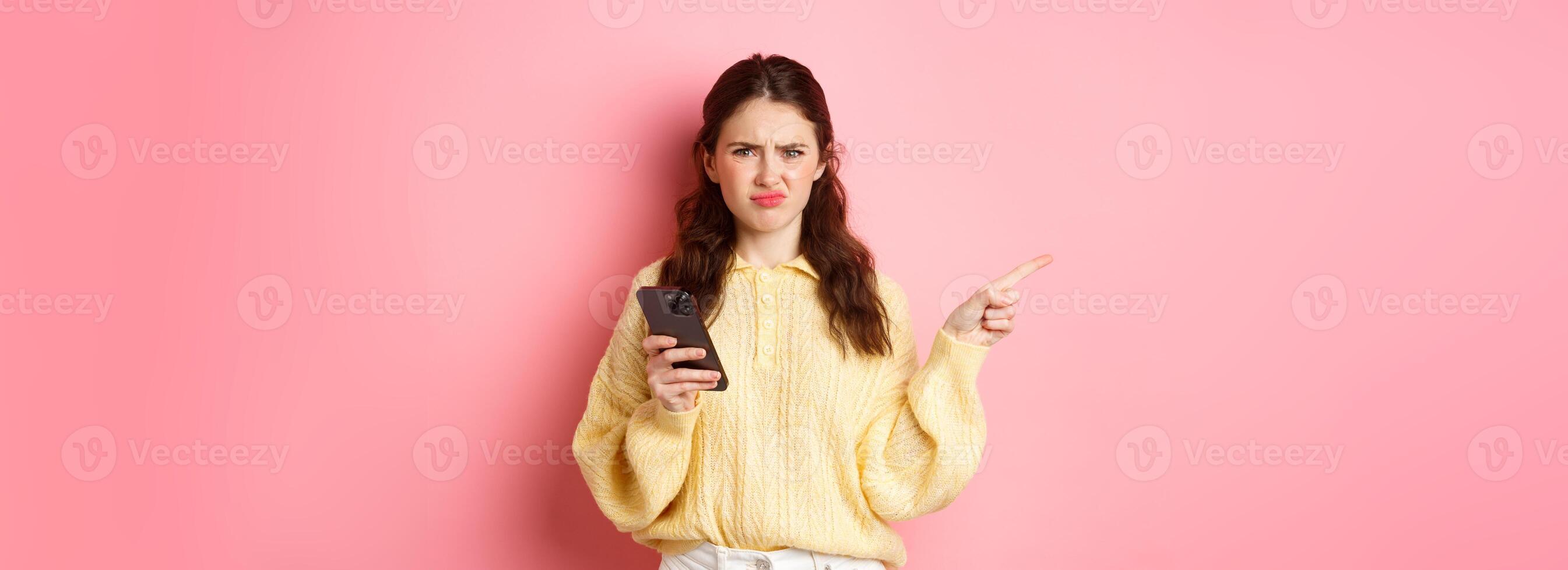 Displeased girl grimacing, frowning upset and pointing finger aside at left copyspace while holding smartphone, standing over pink background photo