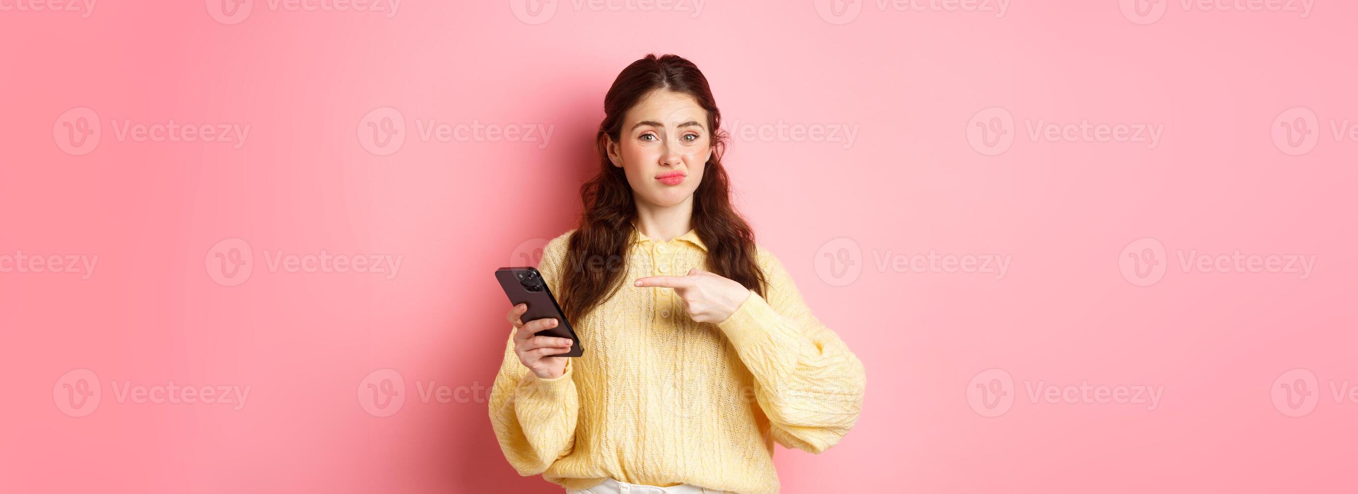 Skeptical young woman grimacing unsatisfied, pointing finger at smartphone screen, having doubts about online content, standing against pink background photo