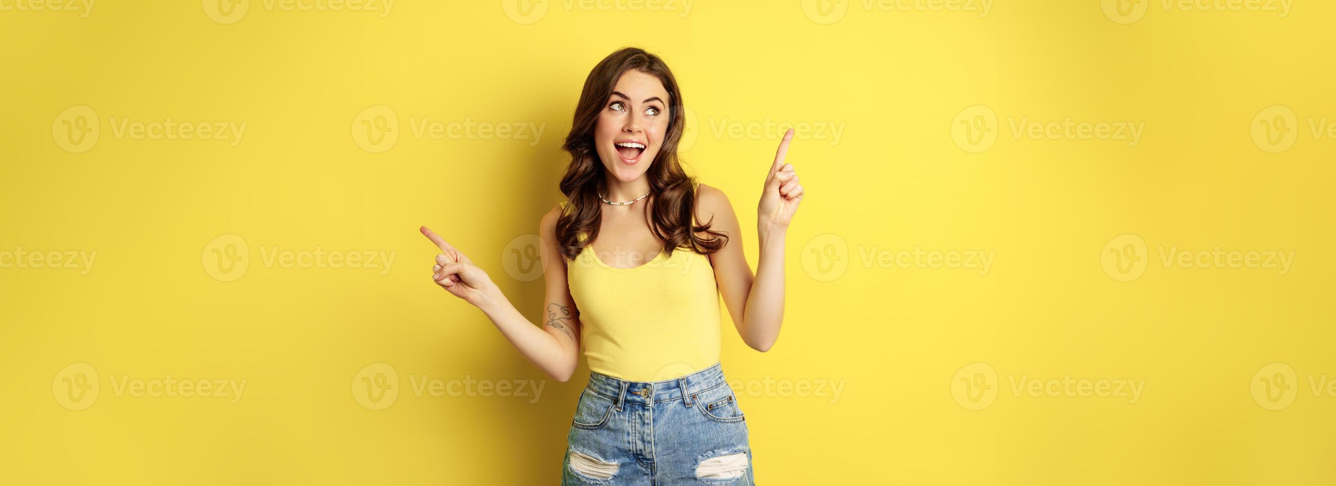 Portrait of stylish young female model, showing advertisement, pointing at logo or banner, demonstrating two ways, choices in store, standing over yellow background photo