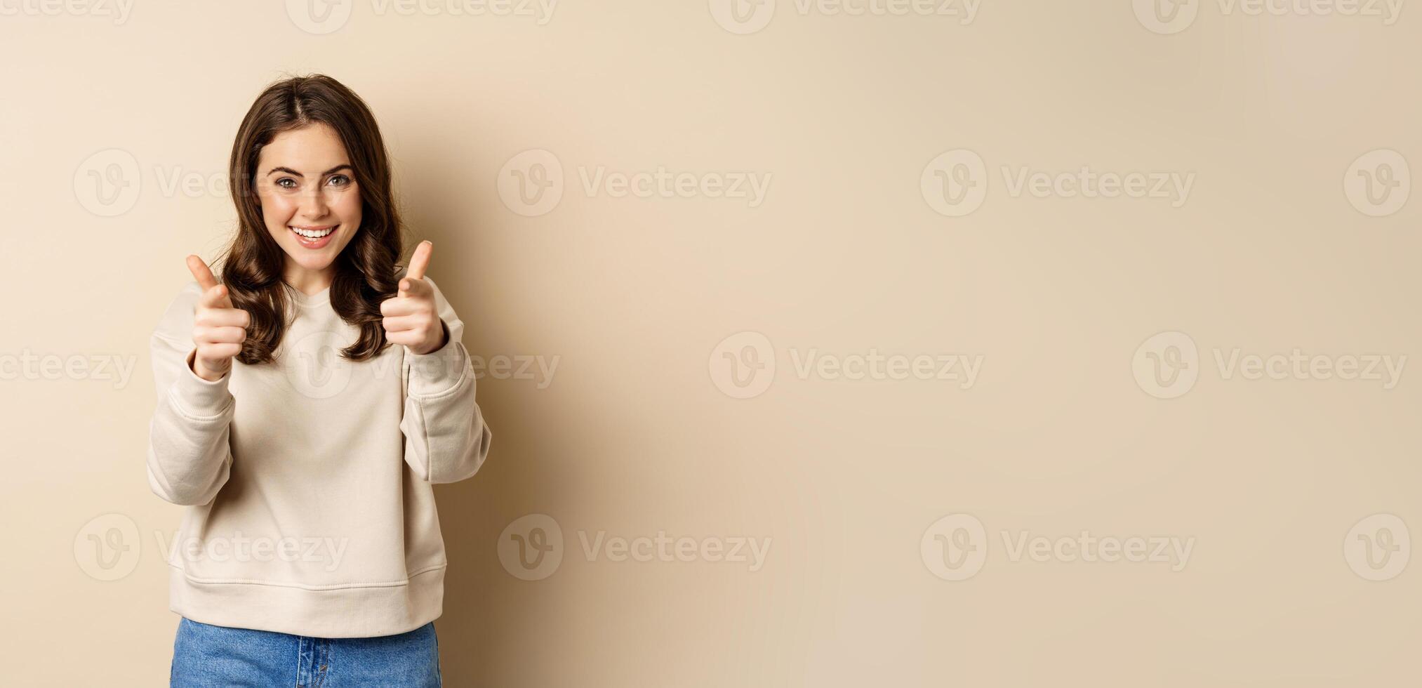 Smiling happy woman choosing you, pointing fingers at camera and laughing, inviting people, standing over beige background photo