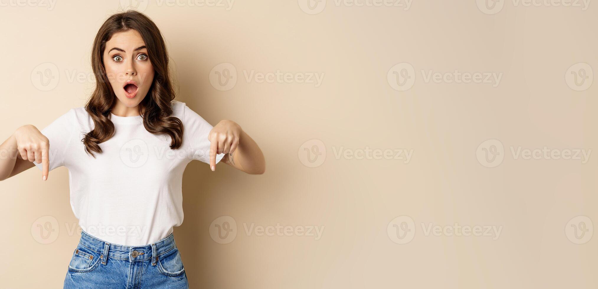 Surprised modern womn pointing fingers down, gasping amazed, showing smth awesome below, standing over beige background photo