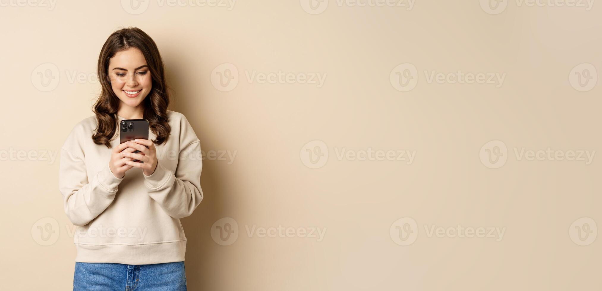 People and cellular technology. Beautiful stylish woman using mobile phone, smartphone app, smiling and looking at screen, standing over beige background photo