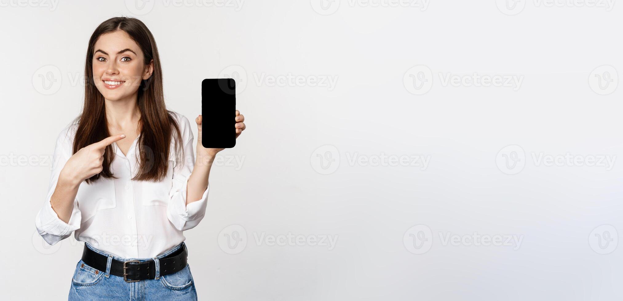 Happy beautiful woman smiling, pointing at mobile phone screen, showing advertisement, website, standing over white background photo