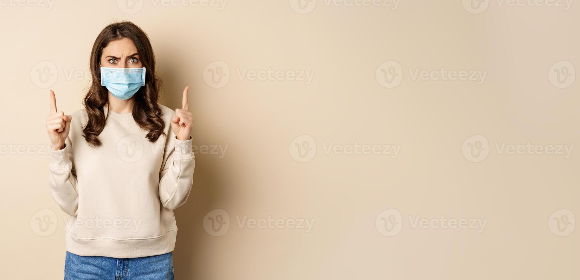 Confused woman in medical mask looking puzzled, pointing fingers up, showing smth strange on top, standing against beige background photo