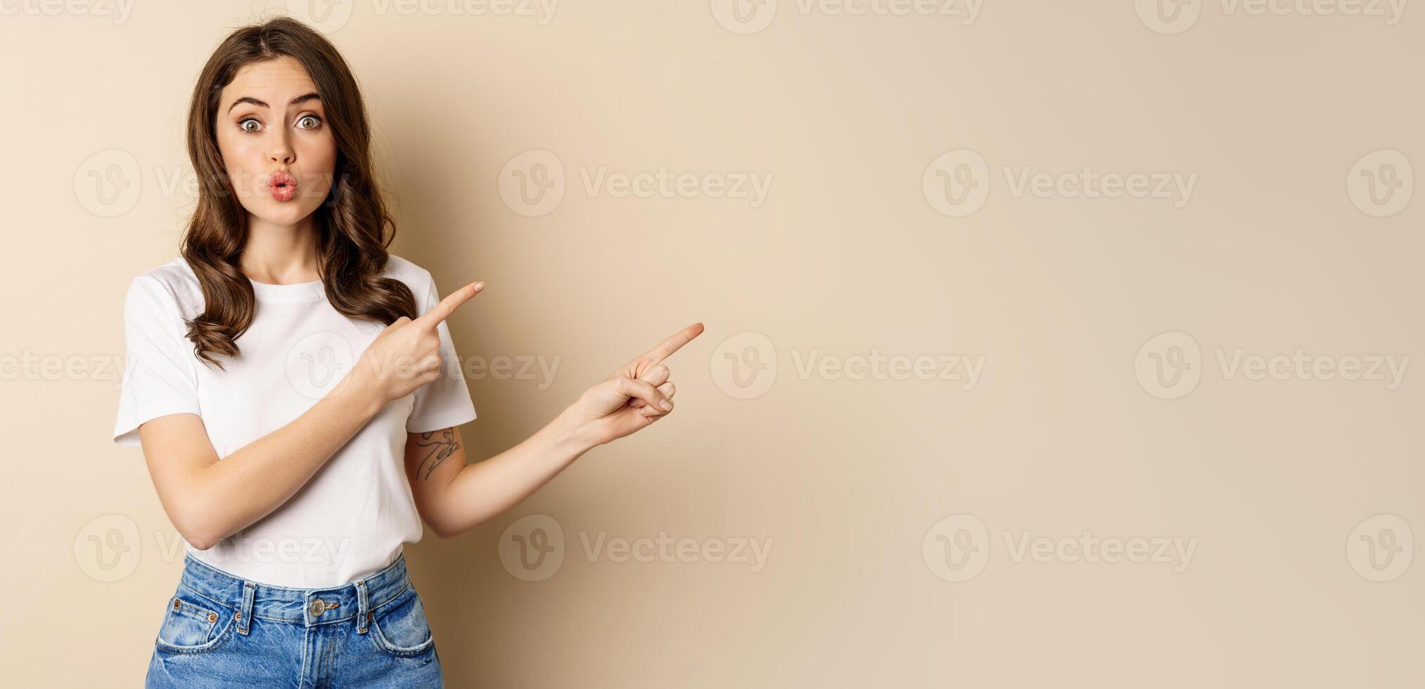 Excited girl pointing fingers right, showing promo with curious face expression, standing over beige background photo