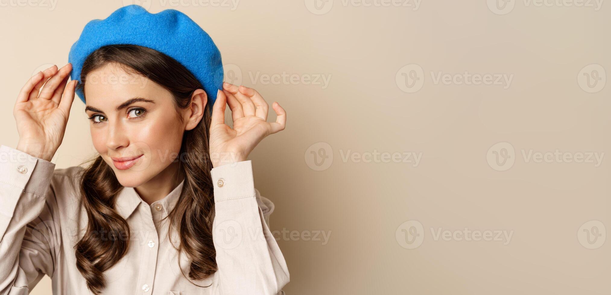 Stylish modern girl put on trendy hat on head and smiling, going out, posing against beige background photo