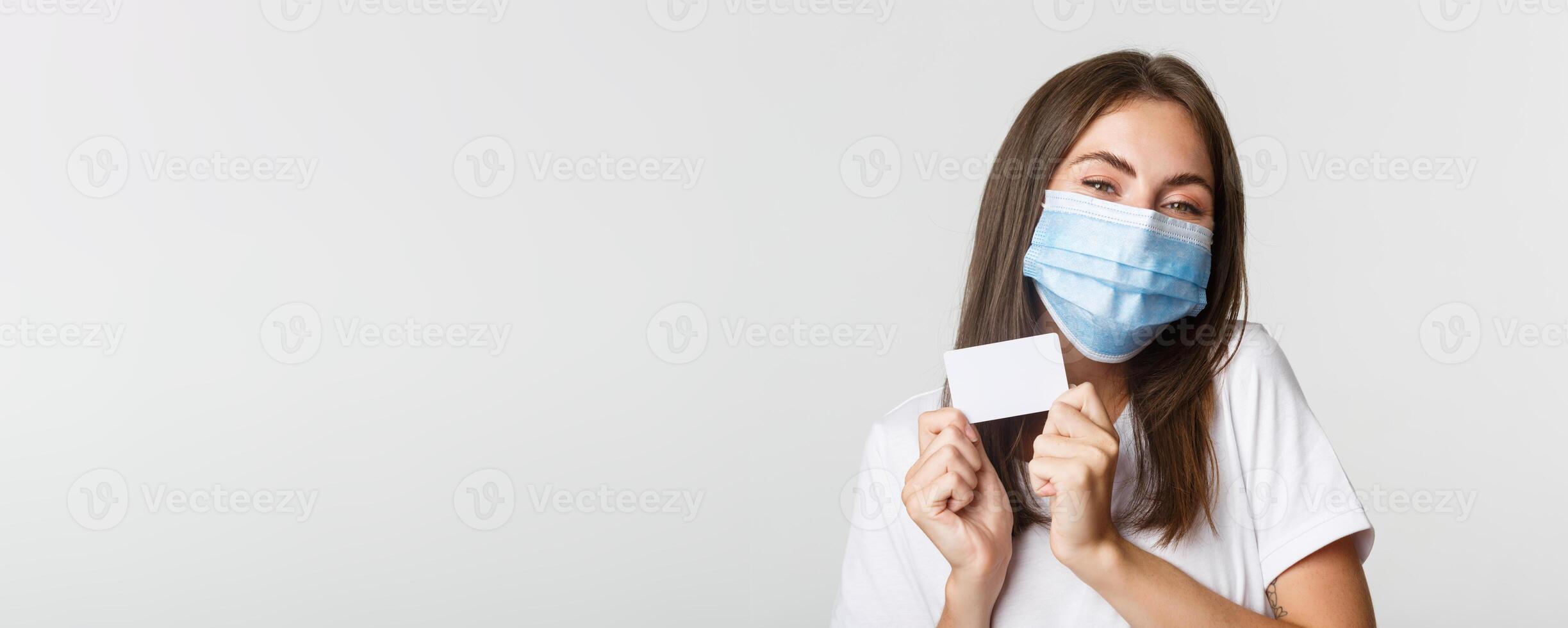 Covid-19, health and social distancing concept. Close-up of attractive happy brunette girl in medical mask showing credit card photo