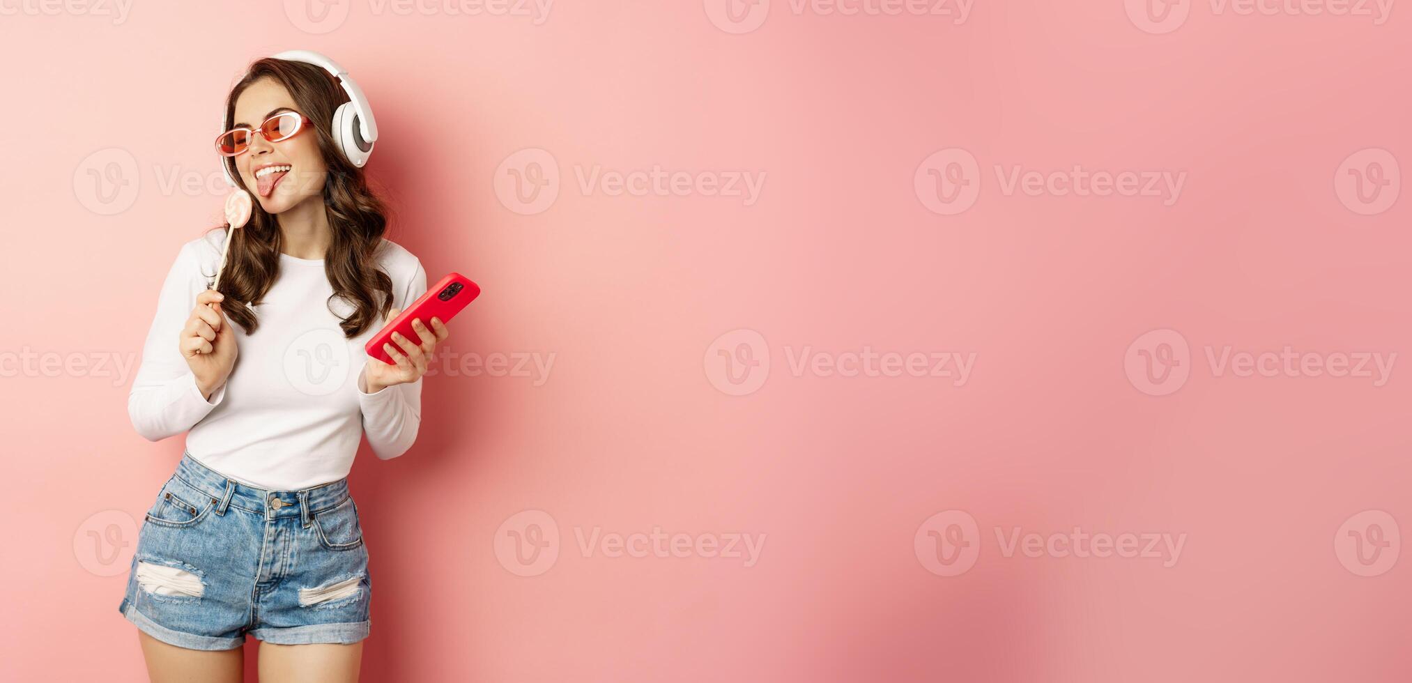 Stylish summer girl dancing with smartphone and lolipop, listening music in headphones, standing in sunglasses against pink background photo