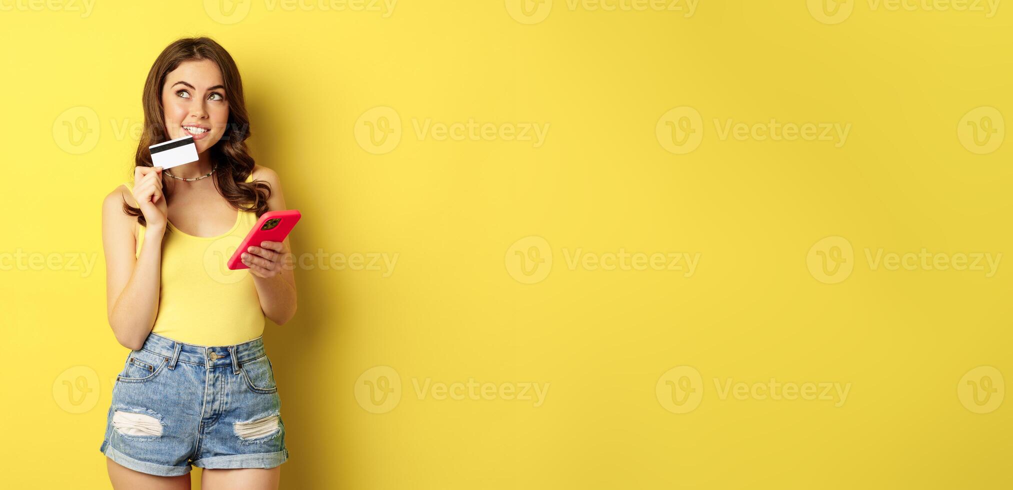 Online shopping. Stylish brunette woman holding smartphone and credit card, paying in app, using mobile phone application, buying smth, standing over yellow background photo