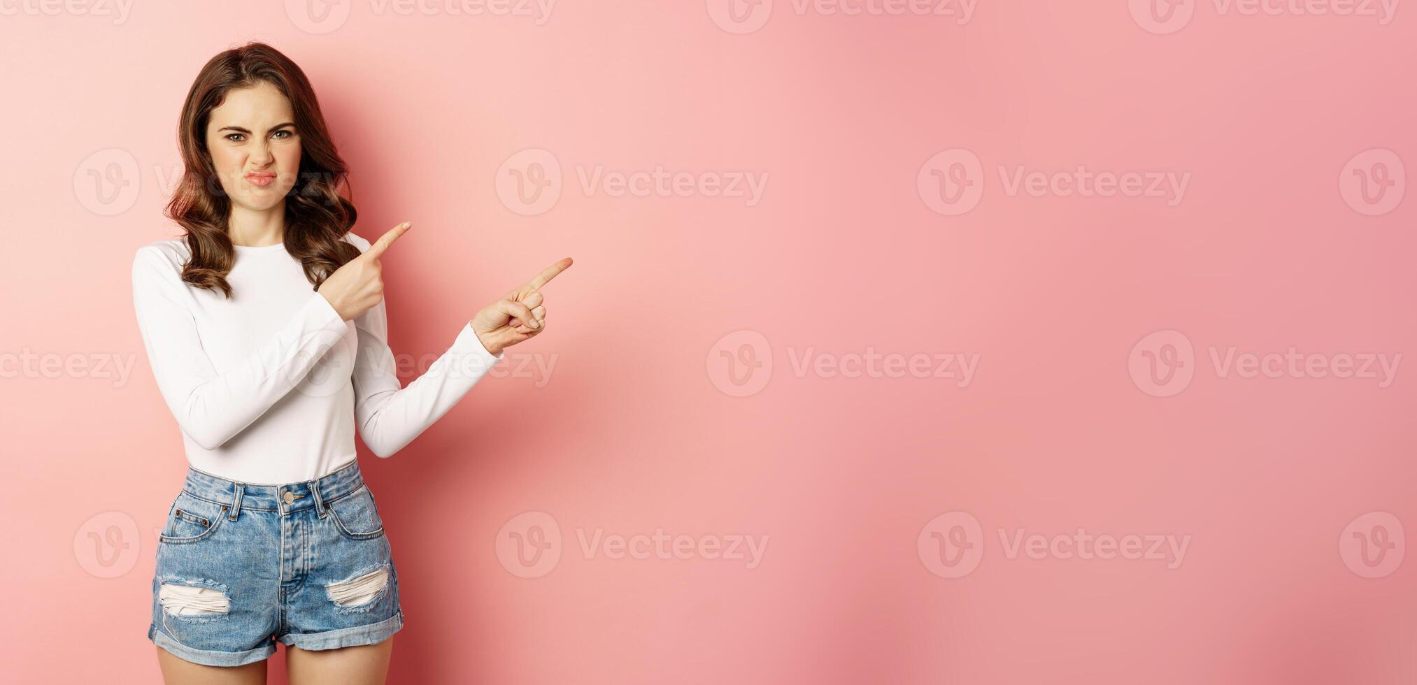 Portrait of attractive girl showing disappointed reaction, pointing fingers right, grimacing from dislike and aversion, complaining at smth bad, pink background photo