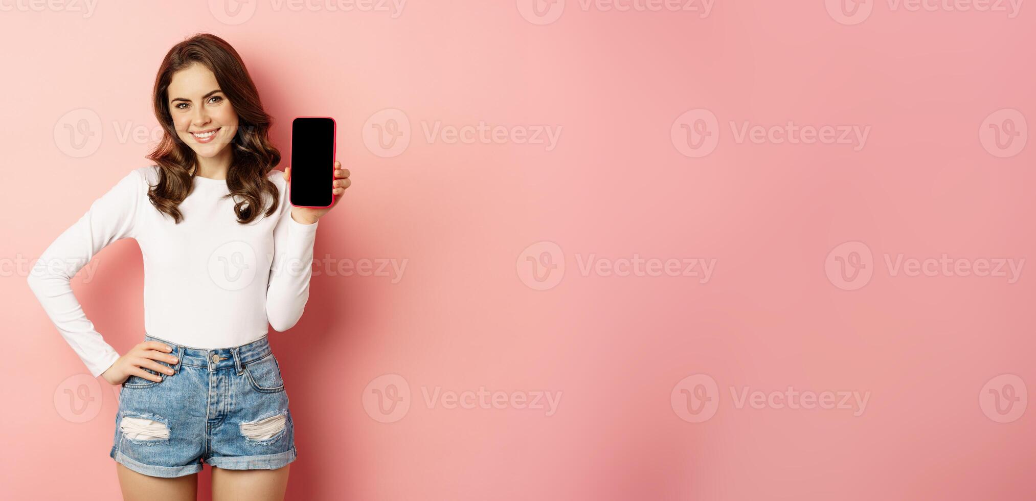 Stylish sexy woman showing mobile phone screen, smartphone app interface and smiling, demonstarting online shop, standing over pink background photo