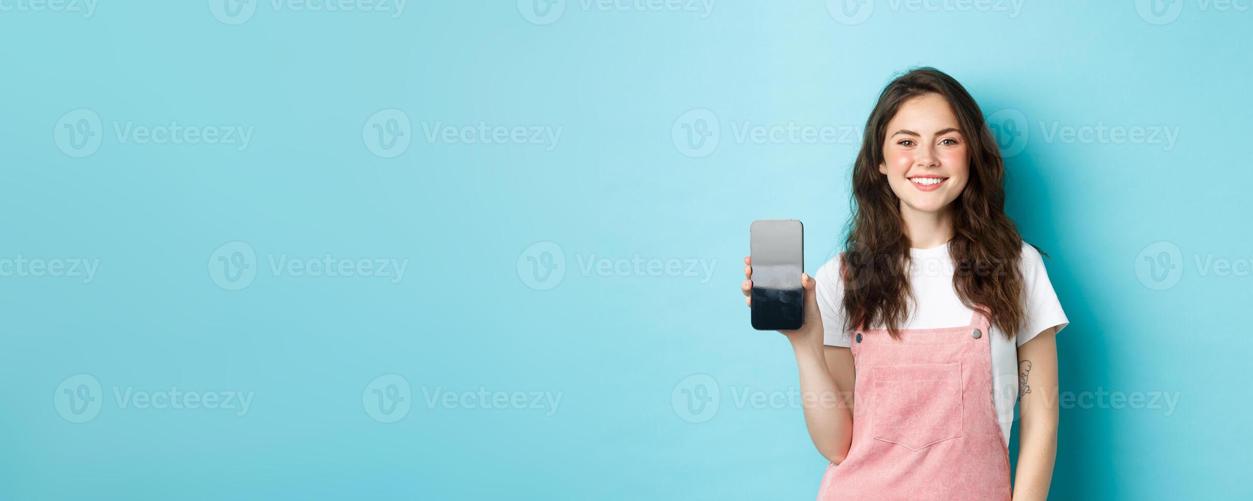 Portrait of smiling beautiful woman showing her smartphone screen and looking pleased, recommending application or online page, standing over blue background photo