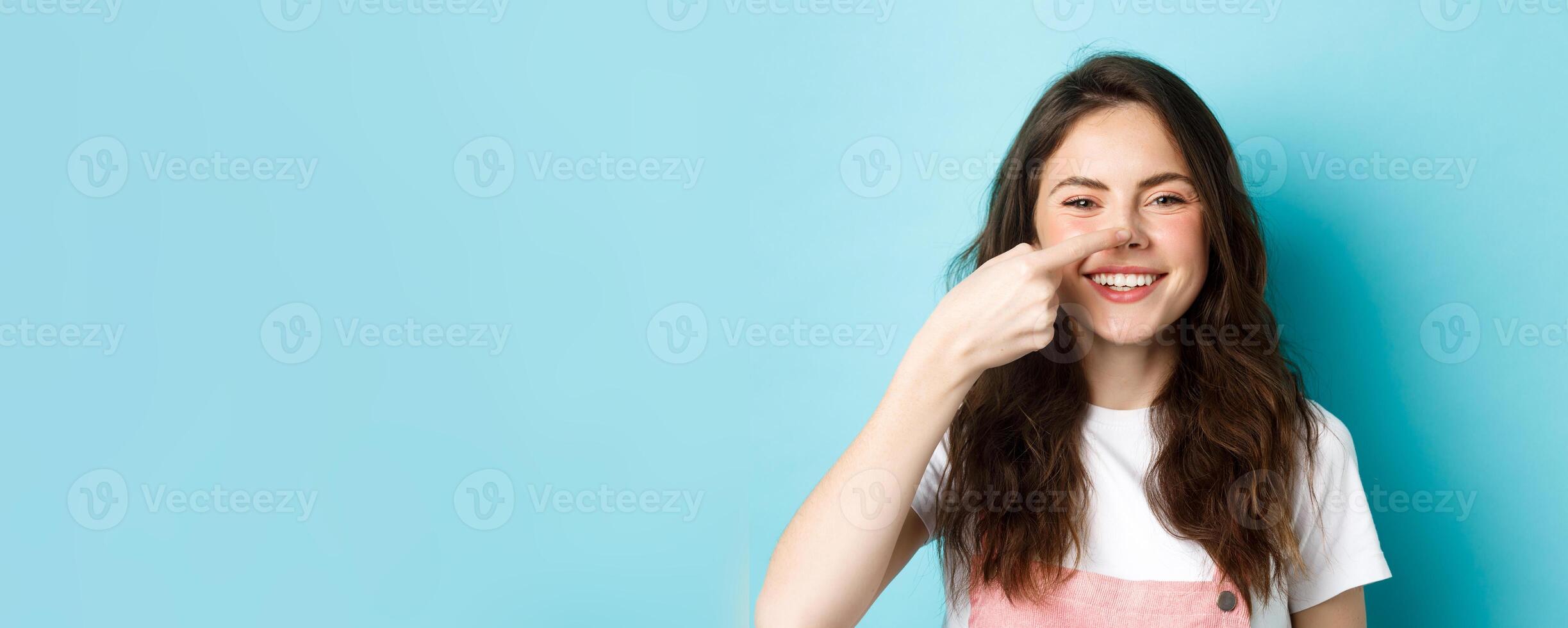 Close up of funny brunette girl touching her nose and smiling cheerfully at camera, standing against blue background photo