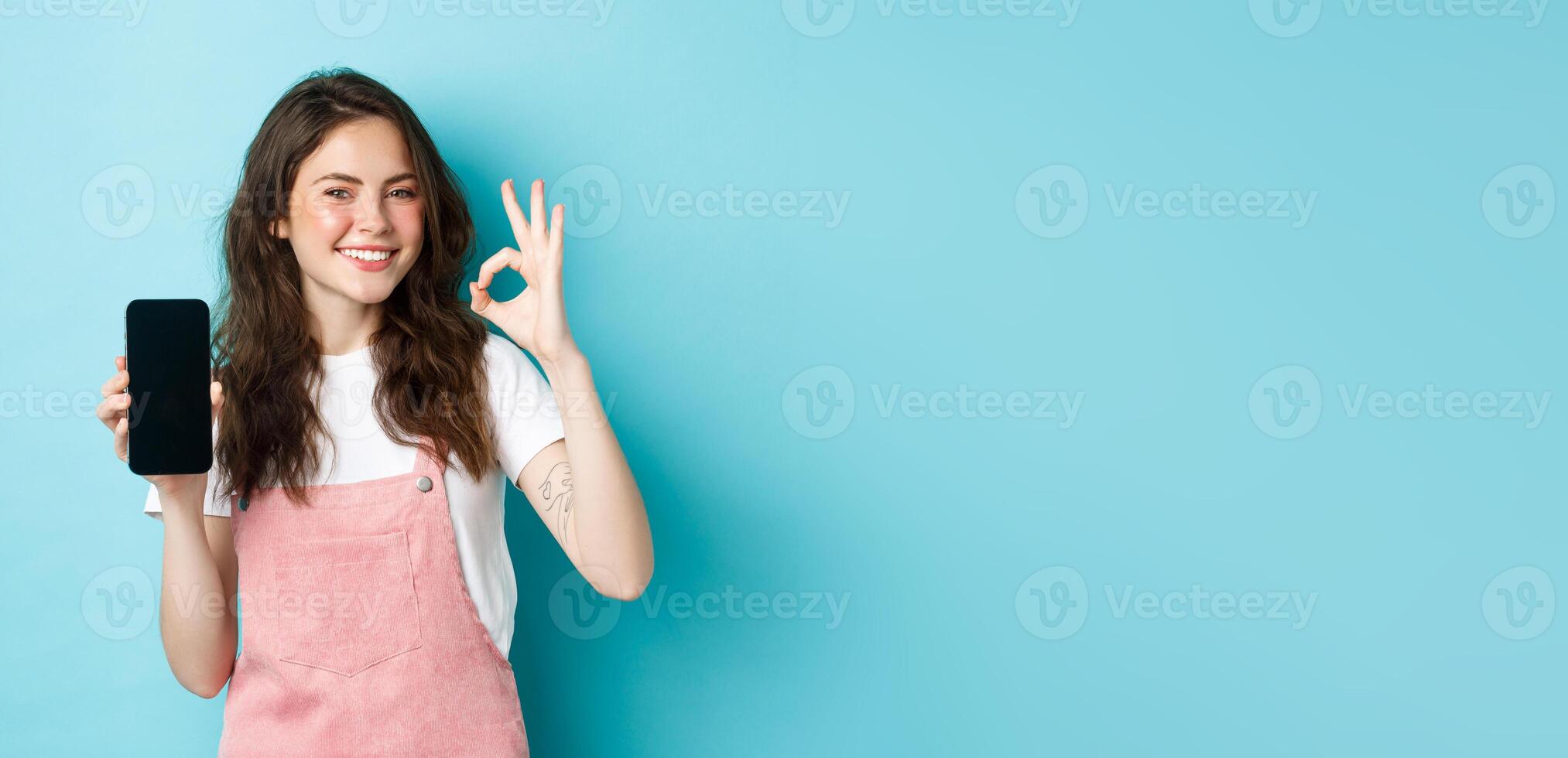 Very good. Smiling young woman showing product online on empty smartphone screen, recommending your brand, make ok sign, standing satisfied against blue background photo