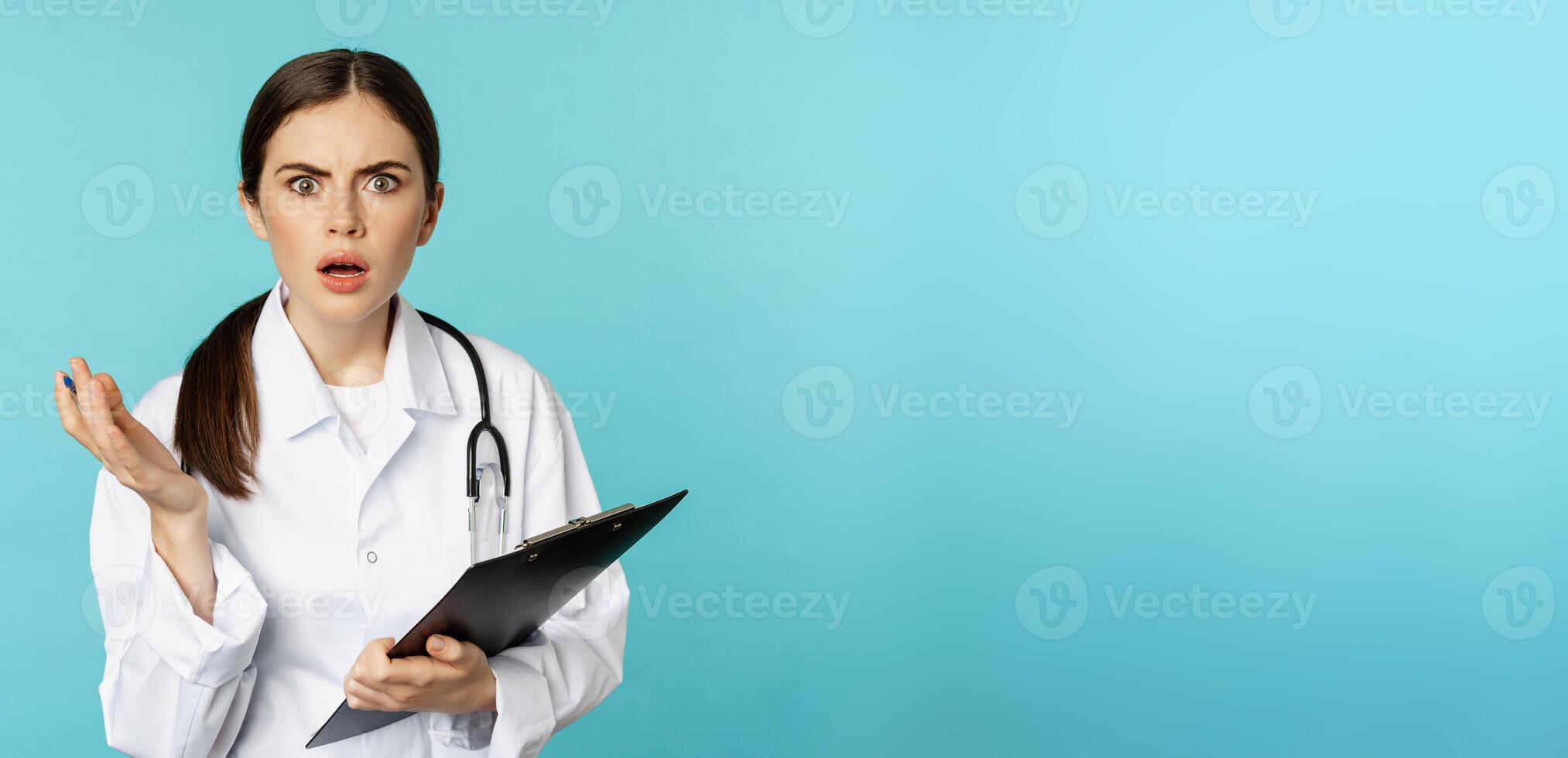 Shocked woman doctor reacting worried and frustrated at camera, hear smth strange, standing in white coat against torquoise background photo