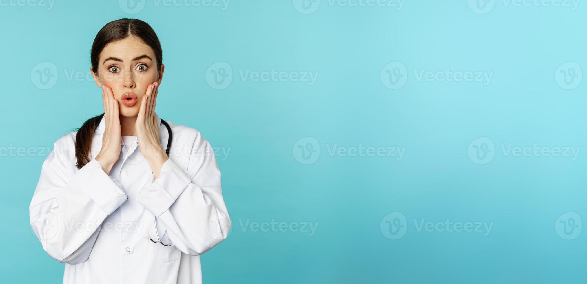 Portrait of shocked female doctor, worried healtcare worker in white robe, looking concerned at patient, standing over blue background photo