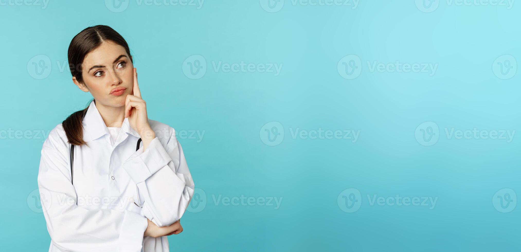 Thoughtful medical worker, young doctor woman looking unamused, bored, standing in white coat against blue background photo