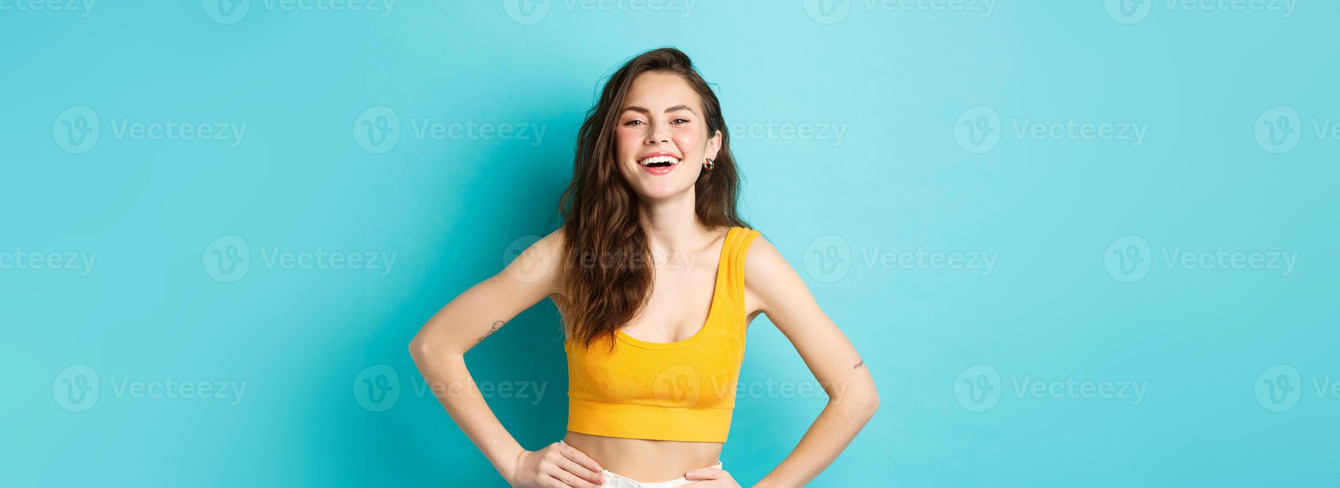 Image of confident and happy stylish woman laughing, holding hands on waist and smiling cheerful, standing carefree in summer outfit, blue background photo