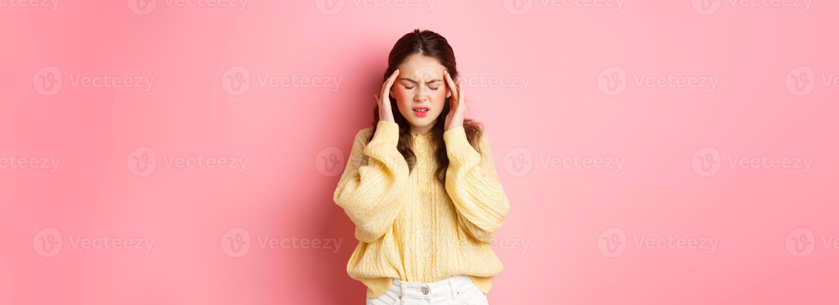 Young woman having terrible headache, touching head and grimacing from pain of migraine, need painkillers, standing against pink background photo