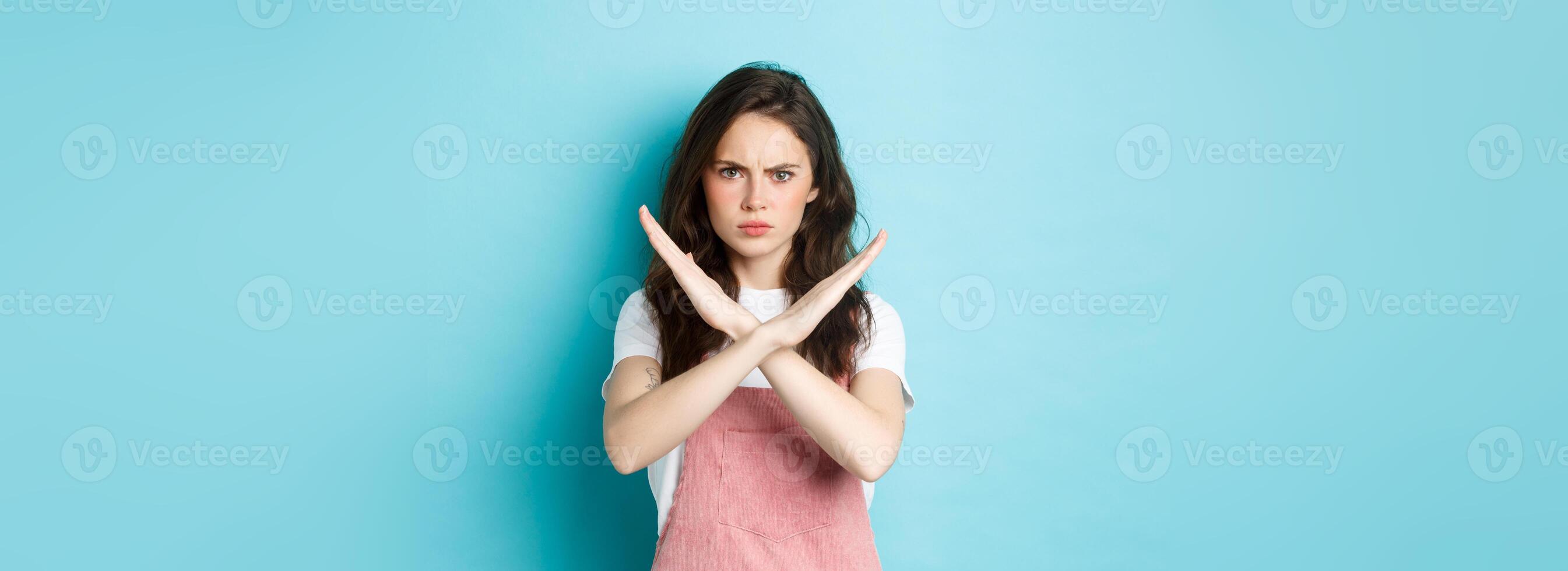 Image of young disappointed woman frowning, making cross sign and sulking, showing stop gesture, say no, forbid bad action, standing over blue background photo